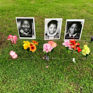Black and white headshots of Carole Robertson, Addie Mae Collins, and Denise McNair, displayed on the lawn with flowers