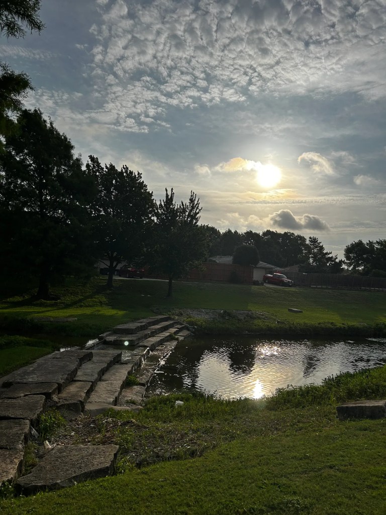 the sun peeking through clouds over a creek with a little bridge made of stones across it and trees along the banks